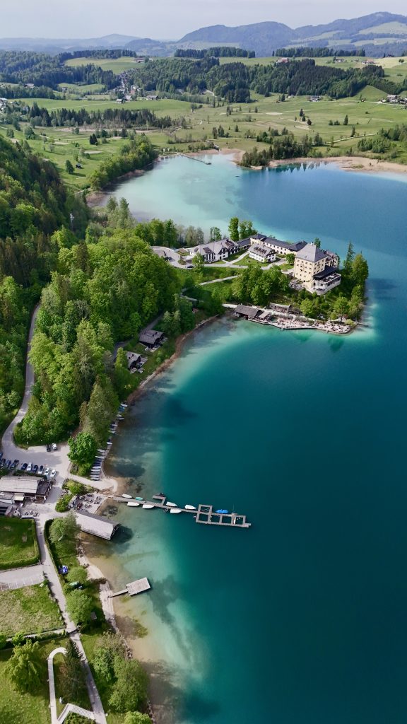 Luftaufnahme eines türkisblauen Sees mit umliegenden Wäldern, Wiesen und einem Hotel am Ufer in einer idyllischen Hügellandschaft.